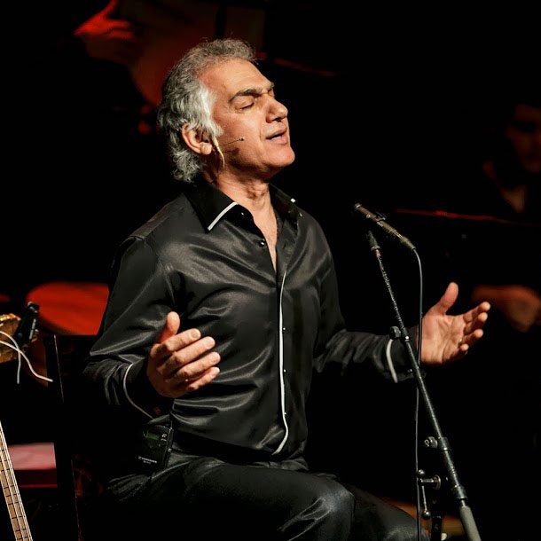 Omar-Faruk-Tekbilek singing on stahe in front of his microphone wearing a black shirt and his hands open, praying