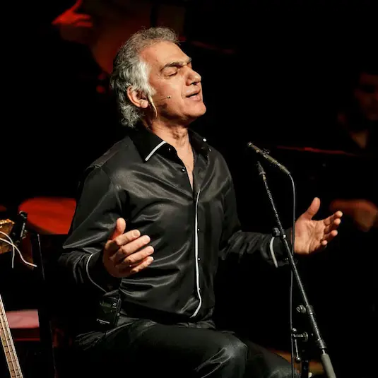 Omar-Faruk-Tekbilek singing on stahe in front of his microphone wearing a black shirt and his hands open, praying